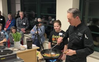 A chef stands at a cooking station with a wok