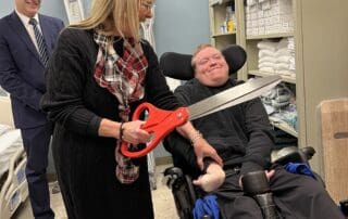 A person seated in a wheelchair indoors while another person beside them holds oversized ceremonial scissors; medical equipment shelves and another standing person are visible in the background.