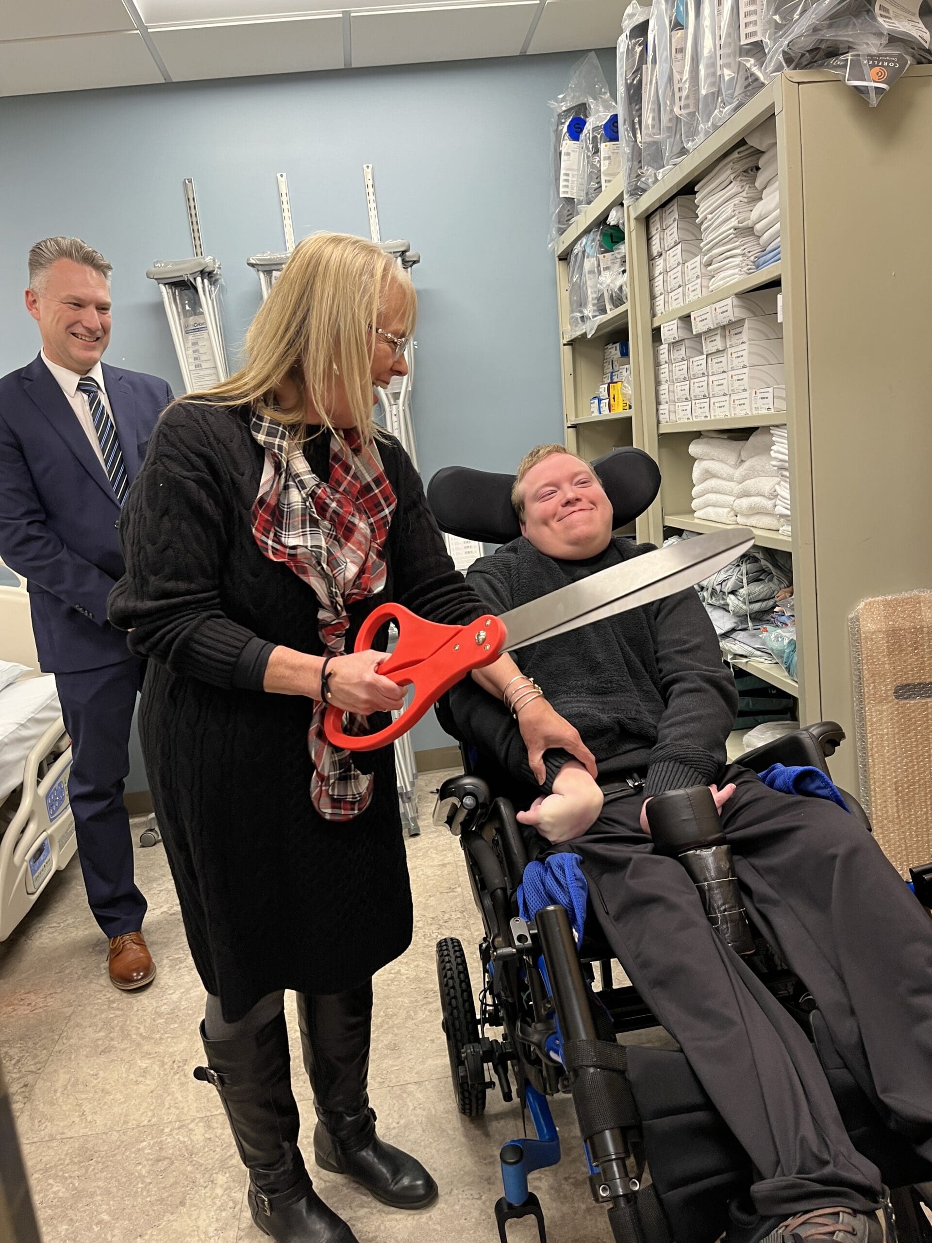 A person seated in a wheelchair indoors while another person beside them holds oversized ceremonial scissors; medical equipment shelves and another standing person are visible in the background.